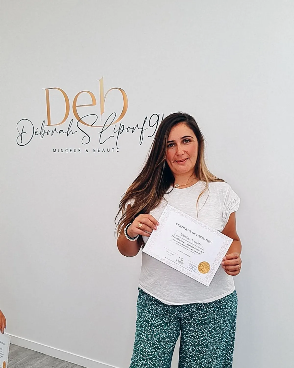 Une stagiaire tient son certificat de formation devant le mur au lettrage doré du centre.